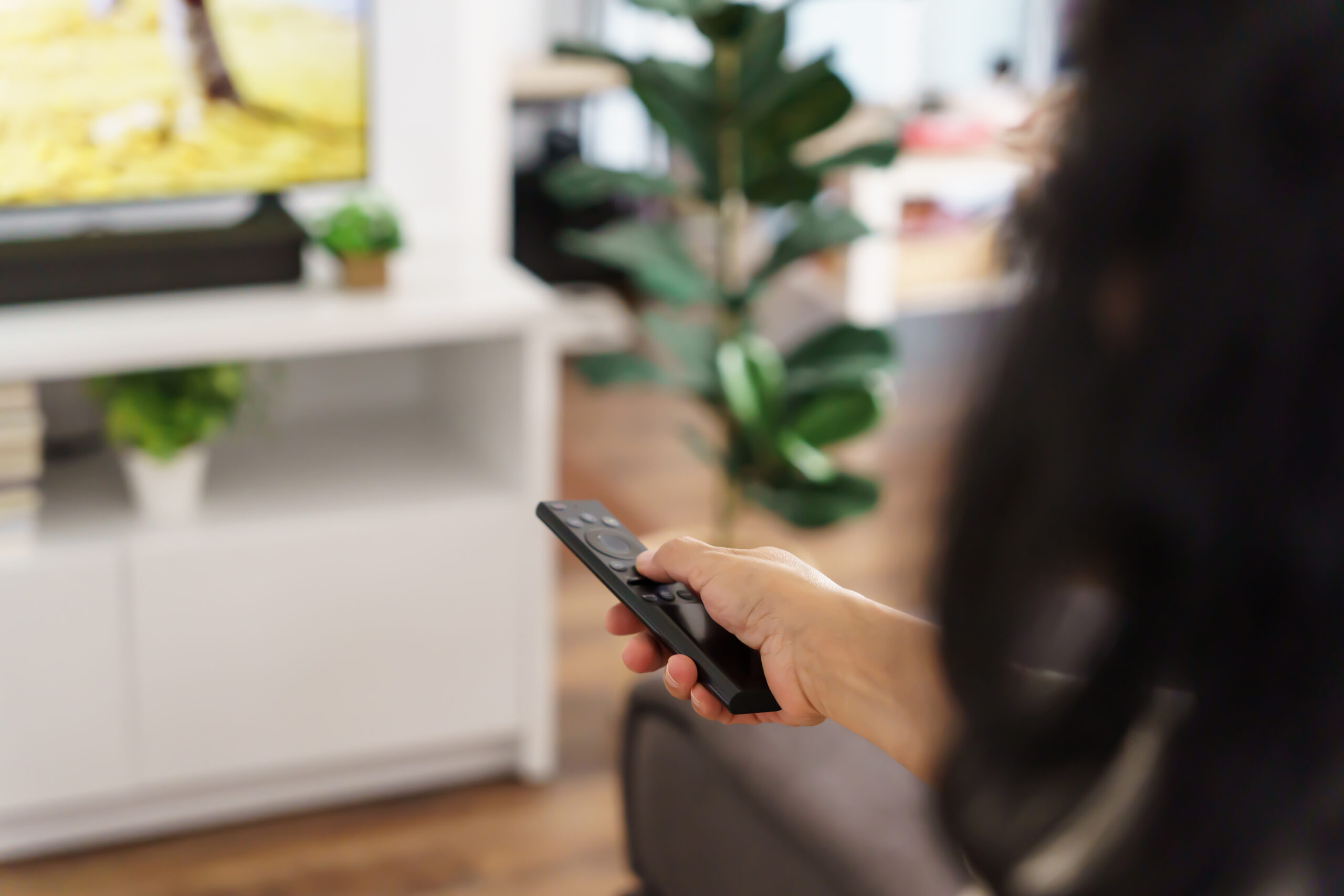 close up of a person's hand holding a remote control for a TV, TV and plan blurred in background.