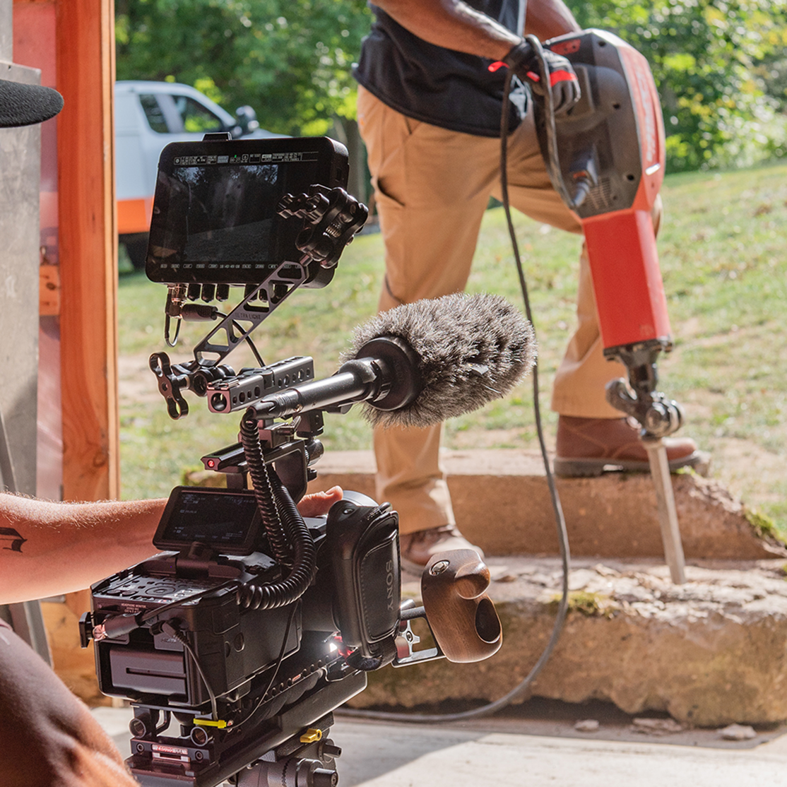 Close-up of a professional video camera rig with a microphone and external monitor, filming a worker using a red jackhammer on concrete steps in the background.