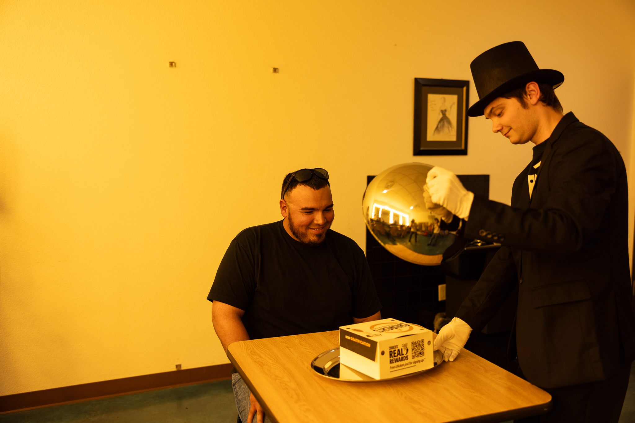 A person dressed in a top hat and white gloves lifts a silver cloche to reveal a box of Church's Chicken to a smiling seated man.