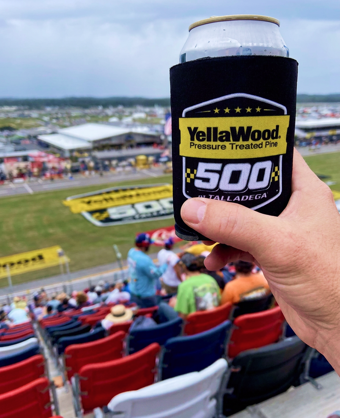 A hand holding a canned beverage in a black "YellaWood 500 at Talladega" koozie, overlooking the grandstands and racetrack in the background.