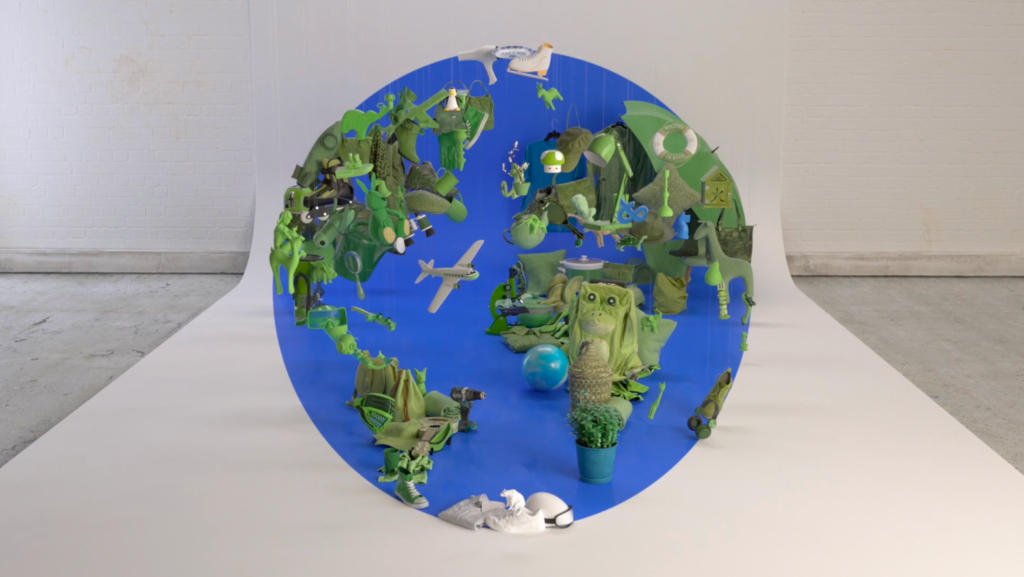 A globe composed of green clothing, toys, and household items arranged as continents against a blue circular background, set in a white photography studio.