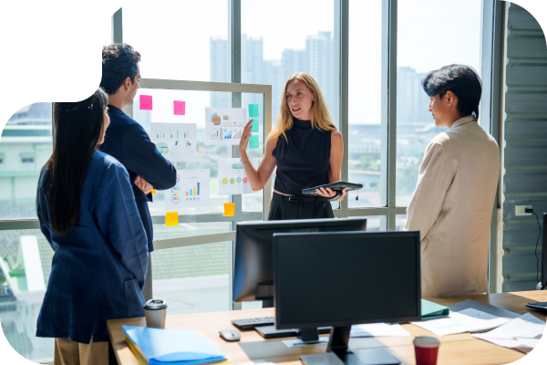 Four people having a stand up meeting in an office in front of a glass board and window