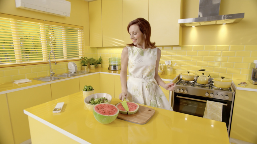 A woman in a white dress slicing a watermelon on the island of a vibrant, entirely yellow kitchen.