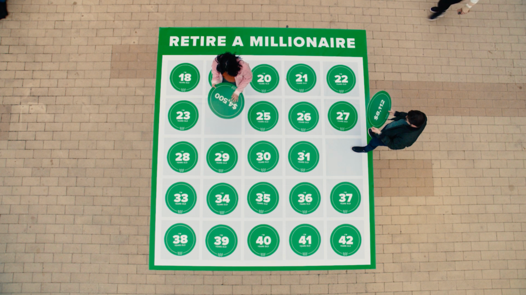 An overhead view of two people interacting with a large floor graphic titled "RETIRE A MILLIONAIRE" that features numbered green circles representing ages.