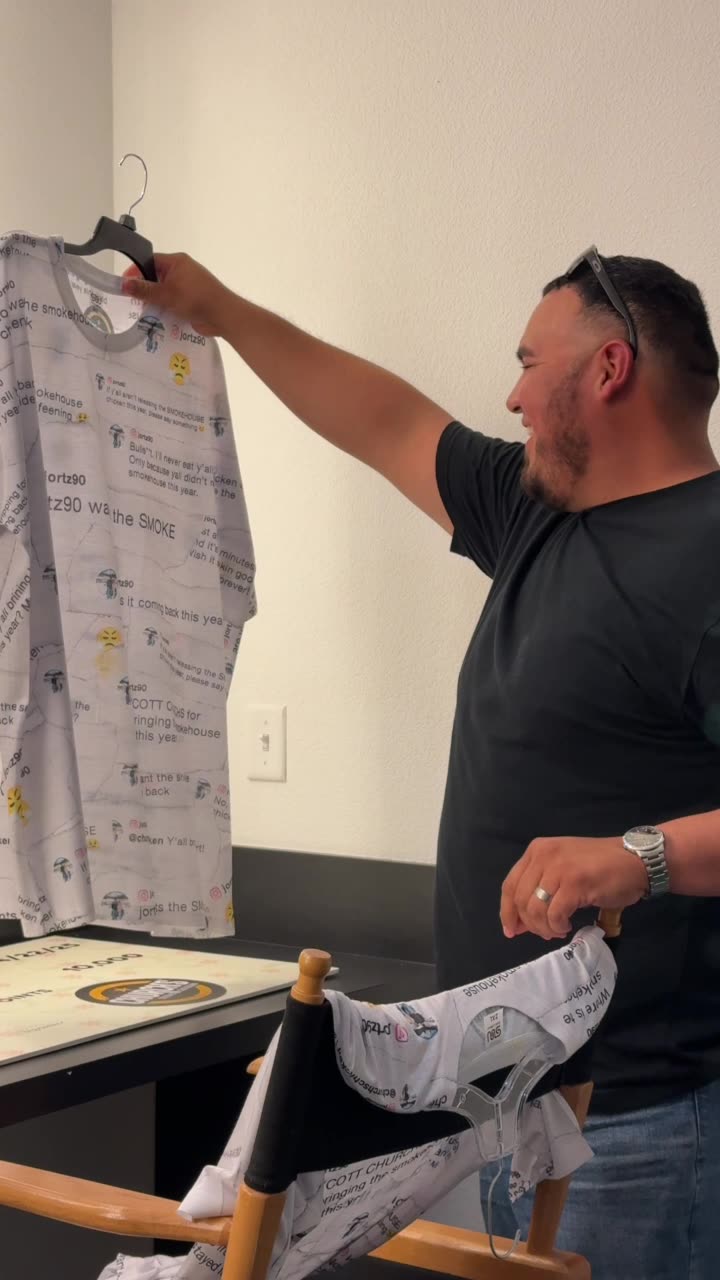 A smiling man holds up one of the custom printed shirts on a hanger, featuring an all-over pattern of social media comments asking for Church's Smokehouse Chicken.